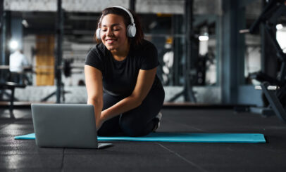 Happy pretty female is sitting on mat and making video training in gym with wireless headphones and notebook